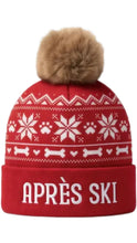 Load image into Gallery viewer, Red après-ski beanie dog toy featuring white snowflake, paw print, heart, and bone patterns, finished with a fluffy brown pom-pom and “APRÈS SKI” lettering on the cuff.