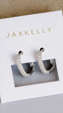 Load image into Gallery viewer, A pair of silver hoop earrings covered in clear pavé stones, presented on a white JAXKELLY brand display card.