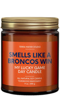 Load image into Gallery viewer, Terra Mater Studio “Smells Like a Broncos Win” 9 oz all-natural soy candle in an amber glass jar with a gold lid, featuring a teakwood mahogany scent and orange-and-navy label.
