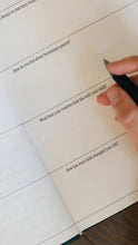 Load image into Gallery viewer, A close-up of a hand holding a black pen and writing in a printed journal or notebook with reflective prompts about becoming a parent.