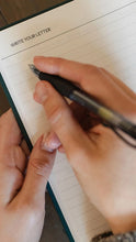 Load image into Gallery viewer, A close-up of a hand holding a black pen and writing on a lined paper letterhead pad, with another hand holding the pad open.