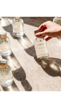 Load image into Gallery viewer, Hand holding a bottle on a textured surface with other bottles in the background.