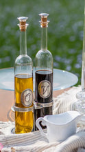 Load image into Gallery viewer, Two clear glass bottles with cork stoppers, one filled with golden oil and the other with vinegar, each featuring a circular monogrammed label, displayed on a tablecloth outdoors with a white pitcher in the foreground.