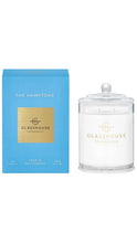 Load image into Gallery viewer, A glass jar candle with a clear lid sits beside its bright blue box. The packaging features a gold square label with the brand name 'GLASSHOUSE FRAGRANCES' and 'THE HAMPTONS' printed above.
