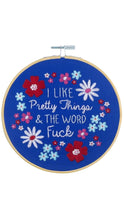 Load image into Gallery viewer, Wooden embroidery hoop featuring bright blue fabric embroidered with colorful red, pink, white, and light blue flowers surrounding the phrase “I Like Pretty Things &amp; The Word Fuck” in white stitching, shown against a clean white background.
