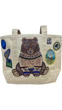 Load image into Gallery viewer, Natural canvas tote bag featuring an embroidered brown bear wearing a patterned knit sweater, surrounded by colorful outdoor-inspired patches, including camping, skiing, and Yosemite National Park designs. Features two sturdy canvas shoulder handles.
