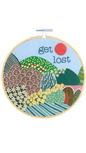 Load image into Gallery viewer, Wooden embroidery hoop featuring a colorful stitched landscape with rolling patterned hills, flowers, and a red sun against a light blue sky, with the phrase “get lost” embroidered in dark blue lettering.
