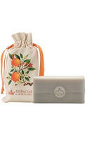 Load image into Gallery viewer, Essências de Portugal bar soap displayed beside a cream drawstring pouch decorated with oranges, green leaves, white blossoms, and orange ribbon ties, featuring the brand name and crown logo, shown against a clean white background.
