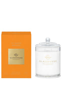 Load image into Gallery viewer, Product photo of Glasshouse Fragrances "St Barts Bronze" soy candle in Vetiver &amp; Grapefruit scent  shown next to its orange packaging box. The candle is in a clear glass jar with a glass lid, featuring a gold "Glasshouse Fragrances" logo on a white label. The matching orange box displays the scent name and a gold square with the same logo. Both items are set against a plain white background.
