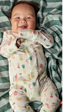 Load image into Gallery viewer, A smiling baby lying on a green and white striped blanket, wearing a cream-colored footed sleeper with a colorful vegetable print.
