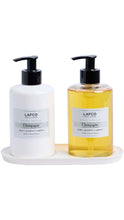 Load image into Gallery viewer, Two LAFCO New York Champagne hand wash bottles on a white oval tray, one white bottle with black pump and one clear bottle filled with golden-yellow liquid, both with white labels.

