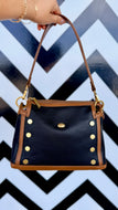A black pebbled leather handbag with tan leather trim and gold-tone hardware, held by a hand against a black and white chevron patterned background.