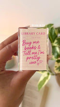 Load image into Gallery viewer, Clear kindle grip with pink text 'Buy me books &amp; Tell me I'm pretty xoxo' held by a hand, with a plant in the background.
