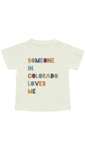Load image into Gallery viewer, A cream-colored short-sleeve baby t-shirt featuring the text 'SOMEONE IN COLORADO LOVES ME' printed on the front in a retro, multi-colored block font.

