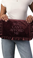 A person holding a deep burgundy velvet clutch bag with ruffled trim and the text 'YOU CAN'T AFFORD ME' in light pink lettering. The person is wearing a white top and blue jeans against a plain white background.