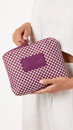 A person in a white dress holds a zippered pouch with a purple and white checkerboard pattern and a purple leather patch on the front. The patch has the brand name 'ALOHA' embossed on it.