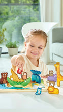 Load image into Gallery viewer, A young child smiling and playing with a colorful wooden animal puzzle toy set on a white table in a bright, modern living room.
