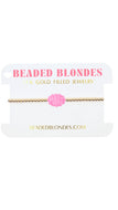 A delicate gold beaded bracelet with a pink monogrammed charm, displayed on a white card labeled 'BEADED BLONDES'.
