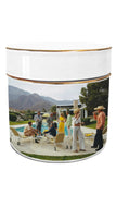 A cylindrical white ceramic tin with a gold-trimmed lid and a printed wraparound photograph of a mid-century modern poolside gathering with mountains in the background.