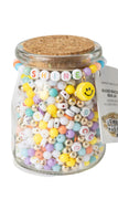 A clear glass jar filled with colorful beaded jewelry, topped with a cork lid and decorated with a beaded 'SHINE' banner and a yellow smiley face charm. A product tag is attached on the right side against a white background.